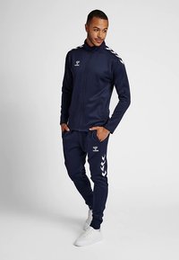 Granatowy dres wykonany z gładkiej tkaniny, z kurtką na zamek błyskawiczny, białym logo i paskami, w komplecie z dopasowanymi spodniami i białymi trampkami.