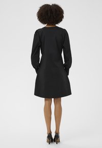 Göndör hajú nő fekete, hosszú ujjú ruhát és fekete magassarkú cipőt visel, háttal áll egy sima háttér előtt.