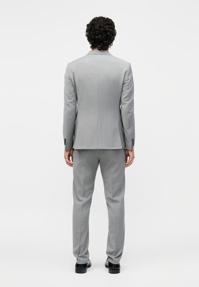 Άνδρας με σγουρά μαλλιά στέκεται με την πλάτη στραμμένη, φορώντας ένα ανοιχτό γκρι κοστούμι και μαύρα επίσημα παπούτσια μπροστά σε απλό λευκό φόντο.