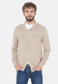Uomo con barba che indossa una camicia beige buttonata sopra una t-shirt bianca e jeans blu, in piedi con le mani in tasca su uno sfondo semplice.