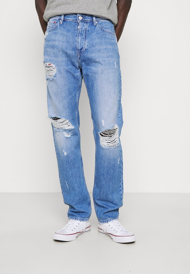 Světle modré džíny s rovně střiženými nohavicemi, které mají velké odřené oblasti, třepící se okraje a mírně výrazný vzhled opotřebení. Nosené s bílými teniskami.