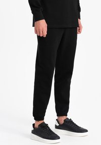 Sorte sweatpants med elastiske manchetter, der har to frontlommer og diskrete syninger. Sammen med sorte sneakers med en hvid sål.