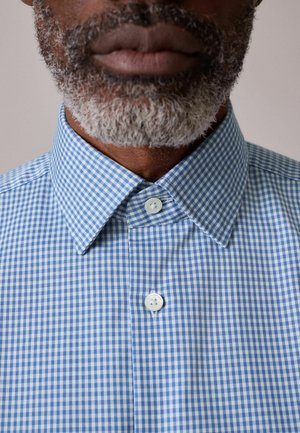 Man with gray beard wearing a blue and white checkered button-up shirt with pointed collar against a plain background.