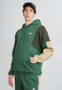 Vihreä ja beige huppari, jossa on kengurupussi, Nike-logo rinnassa ja värikohdistetut hihat. Pehmeä kangas, rento istuvuus.