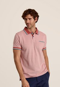 Polo shirt rose clair avec un col rouge, des finitions bleues et vertes sur les manches et la poche. Manches courtes, texture douce et patte de boutonnage.