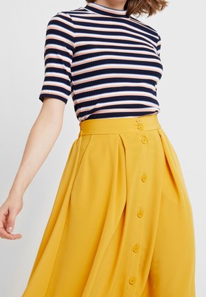 Femme portant un haut côtelé rayé bleu marine, blanc et rose avec des manches au niveau du coude et une jupe jaune boutonnée taille haute.