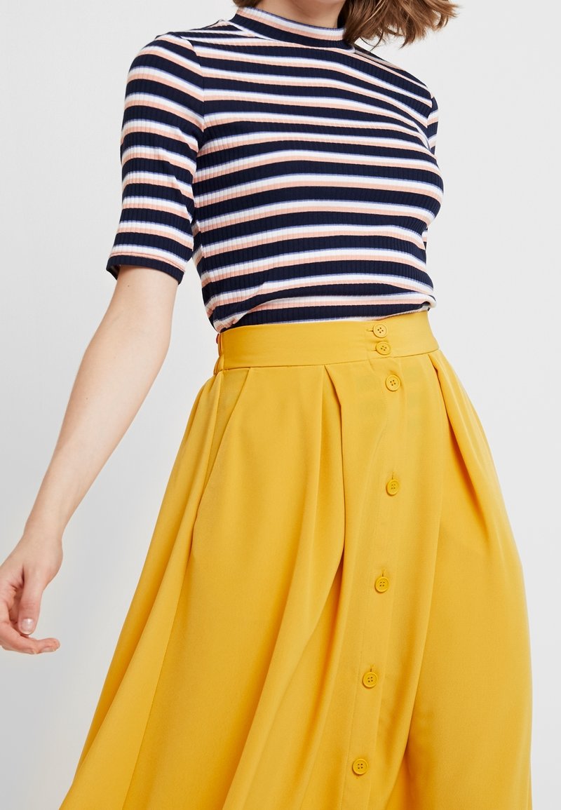 Femme portant un haut côtelé rayé bleu marine, blanc et rose avec des manches au niveau du coude et une jupe jaune boutonnée taille haute.