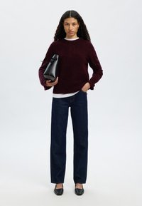 Ogrinjalo iz bordo volne preko bele srajce, v kombinaciji z temno modrimi kavbojkami z ravnimi hlačnicami in črnimi čevlji z ostrim koncem. Model drži majhno črno torbico.