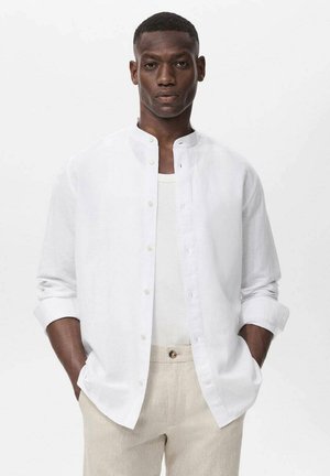 White linen button-up shirt with a band collar, long sleeves rolled at the cuffs, worn over a white tank top, paired with light beige trousers.