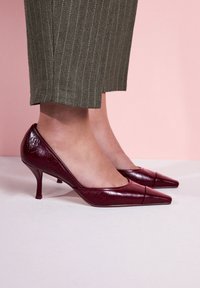 Vínovočervené kožené lodičky na vysokom podpätku so špicatými špičkami, hladkým povrchom a tenkými podpätkami, doplnené o antracitové nohavice s tenkými prúžkami.