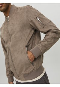 Suede bomberjack in lichtbruin met een ritszak op de linkerarm, ribgebreide manchetten en zoom, en een ritsluiting aan de voorkant. Strakke texture.