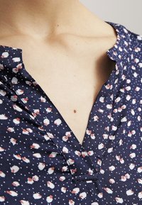 Blusa blu navy con collo a V, presenta un motivo floreale bianco, tessuto morbido e una texture fluida. Vista ravvicinata che mostra i dettagli del colletto.