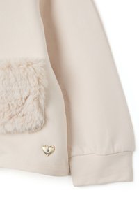 Prenda de manga larga beige con un suave y esponjoso bolsillo de parche y un pequeño adorno metálico dorado en forma de corazón cerca del dobladillo.