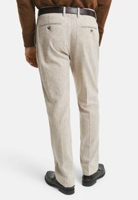 Mann in beigen maßgeschneiderten Hosen mit geknöpften Gesäßtaschen, dunklem Gürtel, braunem Pullover und schwarzen Slippern, von hinten gezeigt.