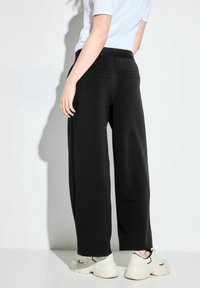 Černé kalhoty širokého střihu vyrobené z měkké tkaniny, s elastickým pasem a detaily kapes, spárované se světlými teniskami.