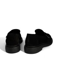 Zwarte suede instappers met een gestructureerde zwarte rubberen zool. Heeft een ronde neus en een decoratieve gespdetail aan één kant.