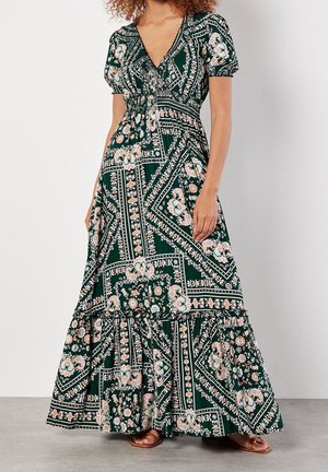 Kvinde iført en mørkegrøn maxikjole med blomsterprint, puffærmer, V-udskæring og flæsekant, kombineret med brune åbne tåsandaler.