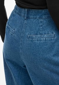 Plave traperice visokog struka s glatkom teksturom, koje imaju dva stražnja džepa i prilagođeni kroj.