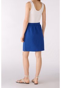 Femme portant un haut blanc sans manches, une jupe bleue au genou avec une fente latérale, et des sandales plates beiges à lacets, debout devant un fond uni.
