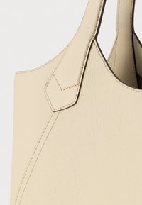 Bēša ādas soma ar fakturētu virsmu, pastiprinātu šuvju un dubulta roktura detaļu, kas redzama uz vienkrāsaina fona.