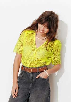 Femme souriante, portant un chemisier en dentelle jaune fluo, ceinture marron, jean gris, collier et bracelet en perles, une main dans la poche.