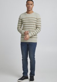 Lichtbeige crewneck trui met bruine horizontale strepen, gecombineerd met blauwe spijkerbroek en zwarte sneakers, gepresenteerd op een model.