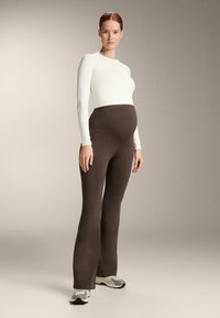 Těhotenský outfit se skládá z bílé dlouhé mikiny s dlouhým rukávem a hnědých rozšířených legín s bočním detailem, navržený pro pohodlí a pohyblivost.