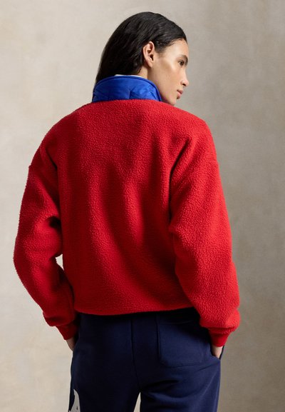 Γυναίκα με σκούρα μαλλιά που φοράει κόκκινο φλις μπουφάν και μπλε παντελόνι, γυρισμένη στο πλάι με τα χέρια στις τσέπες, μπροστά σε απλό ουδέτερο φόντο.