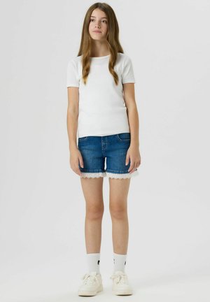 Fiatal lány áll, fehér rövid ujjú ingben, csipkés szegélyű farmer sortban, fehér zokniban és fehér tornacipőben.