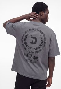 Muž s tmavou pokožkou a krátkymi kučeravými vlasmi nosí sivé oversize tričko s čiernou kruhovou grafikou "DROPSIZE" a "HUSTLE CLUB" na chrbte.
