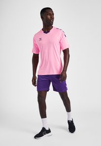 Rosa Sporttrikot mit rundem Ausschnitt, lila Akzenten an den Ärmeln, kombiniert mit lila Shorts. Schwarze Sportschuhe und weiße Socken runden das Outfit ab.