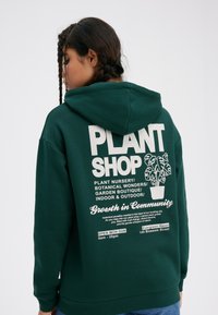 Green hoodie featuring a printed design on the back with text and a plant graphic. Material appears soft; the fit is relaxed.