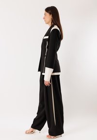 Donna con blazer su misura bianco e nero e pantaloni a gamba larga in piedi di profilo su sfondo bianco neutro.