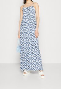 Niebieska i biała, wzorzysta sukienka maxi z dopasowanym gorsetem, cienkimi ramiączkami oraz warstwową spódnicą. Połączona z białymi butami na platformie oraz jasnoniebieską torebką.