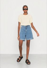 Top curto de manga curta com textura amarelo pálido, calções de ganga de cintura alta com a bainha virada, sandálias pretas e óculos de sol escuros.