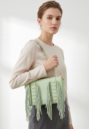 Jonge vrouw met een beige top houdt een lichtgroene handtas met lange franjes over haar schouder tegen een effen achtergrond.