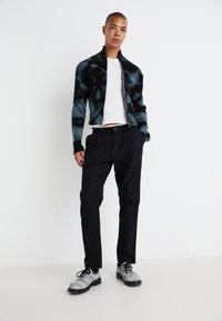 Svart ribbet jakke med blå tie-dye mønster, hvit t-skjorte, svarte bukser og grå sko med svarte snører, stående mot en hvit bakgrunn.