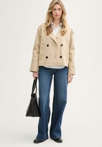 Beige doppelt knopfbares Jacket mit schwarzen Knöpfen, zwei Taschen vorne, getragen über einem weißen Hemd und blauen Schlagjeans. Schwarze Handtasche in der Hand.