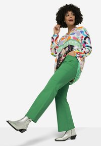 Camisa de gran tamaño multicolor con mangas largas y estampado gráfico, combinada con pantalones rectos verdes y botas de tobillo plateadas.