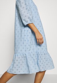 Femme portant une robe bleu clair avec des motifs floraux en eyelet, des manches bouffantes et un ourlet froncé marchant devant un fond uni.