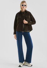 Braune Cordjacke mit zwei Brusttaschen, kombiniert mit blauen weiten Jeans und weißen Sneakers, vor einem neutralen Hintergrund.