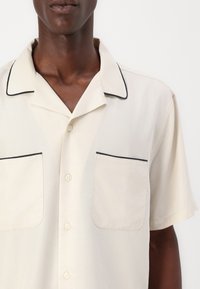 Short-sleeve shirt in light beige fabric with black piping on the collar and pockets. Features two buttoned chest pockets.
