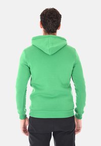 Uomo di spalle che indossa un felpa verde brillante e pantaloni neri su uno sfondo grigio chiaro uniforme.