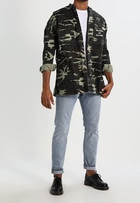 Camicia mimetica verde scuro e nero, con due tasche frontali e uno slogan bianco. Abbinata a jeans azzurri e scarpe nere.