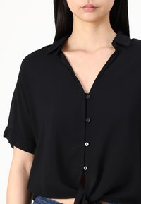 Black short-sleeve blouse made of lightweight fabric, featuring a V-neck, button-down front, and a tied hem detail.