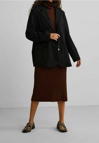 Schwarzer Blazer über einem braunen, gerippten Rollkragenkleid getragen, kombiniert mit gemusterten Slippern. Der Blazer hat einen einzelnen Knopf und Taschen.