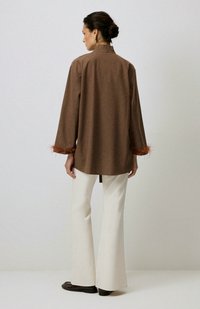 Camicia marrone a maniche lunghe con colletto alto, caratterizzata da dettagli in piuma arancione ai polsini, abbinata a pantaloni bianchi svasati e scarpe scure.