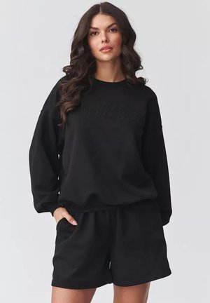 Kobieta o długich ciemnych włosach, ubrana w czarną, za dużą bluzę i pasujące czarne szorty, stoi z jedną ręką w kieszeni.