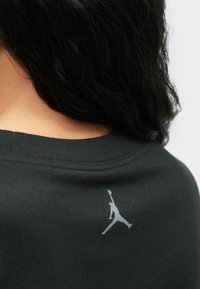 Άτομο με μαύρα μαλλιά που φοράει ένα σκούρο πουκάμισο με ένα μικρό λευκό λογότυπο Jumpman στο επάνω μέρος της πλάτης, κοντά στη λαιμόκοψη.