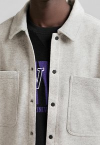 Jasnoszara kurtka koszulowa z mieszanki wełny, z kołnierzem szpiczastym, zatrzaskami i dwoma przednimi kieszeniami, noszona na czarnej bluzie z fioletowymi literami.